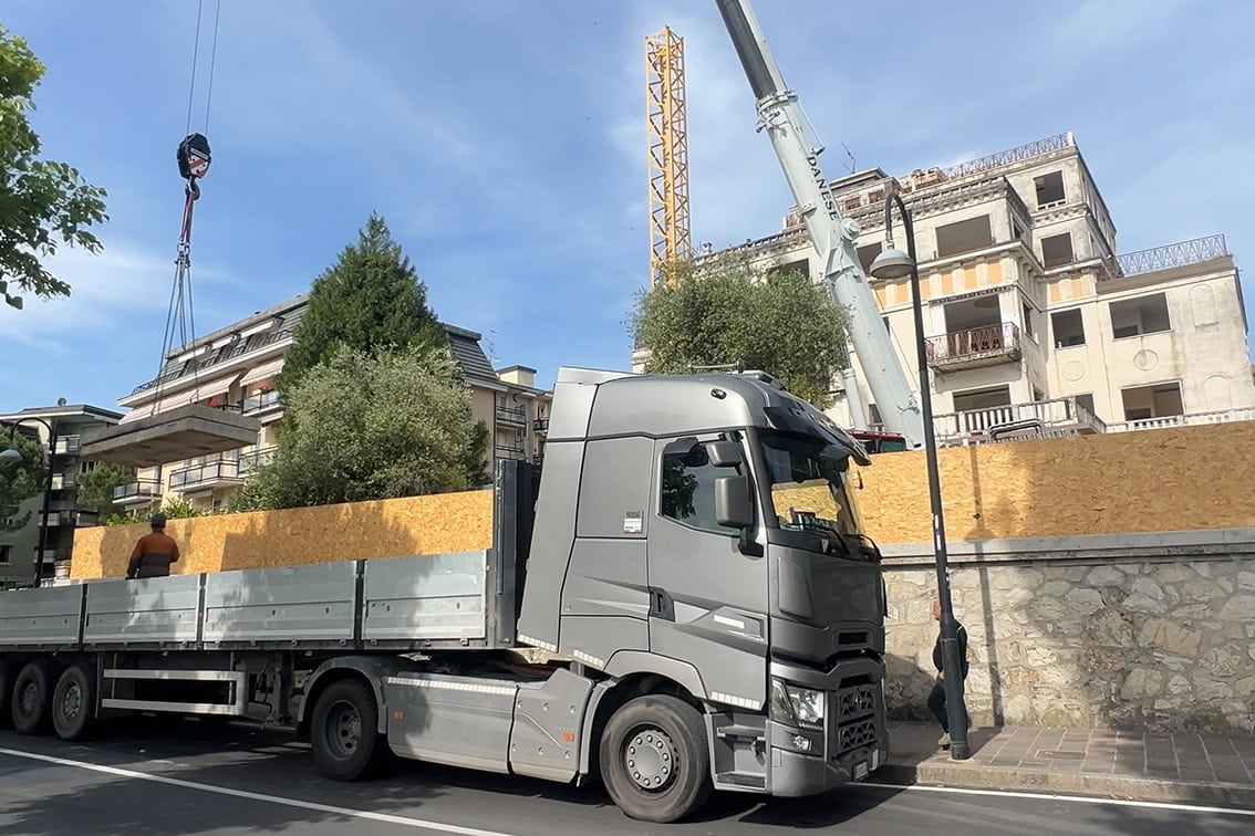 Camion carico di materiali edili in sosta, con operazioni di sollevamento in corso sulla strada pubblica, cantiere Villa Olympia Residences
